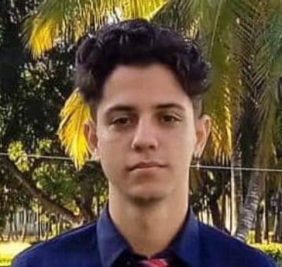 Eduardo is a young Cuban man with dark hair. His head and shoulders are visible and he is wearing a blue shirt. He is looking directly at the camera and standing in front of some palm trees