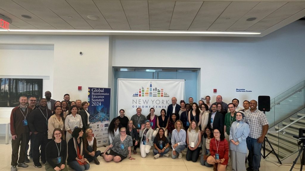 A group of people gathered in front of a sign that says "New York Genome Center". The the left is a banner saying "Global Bioinformatics Education Summit 2024". 