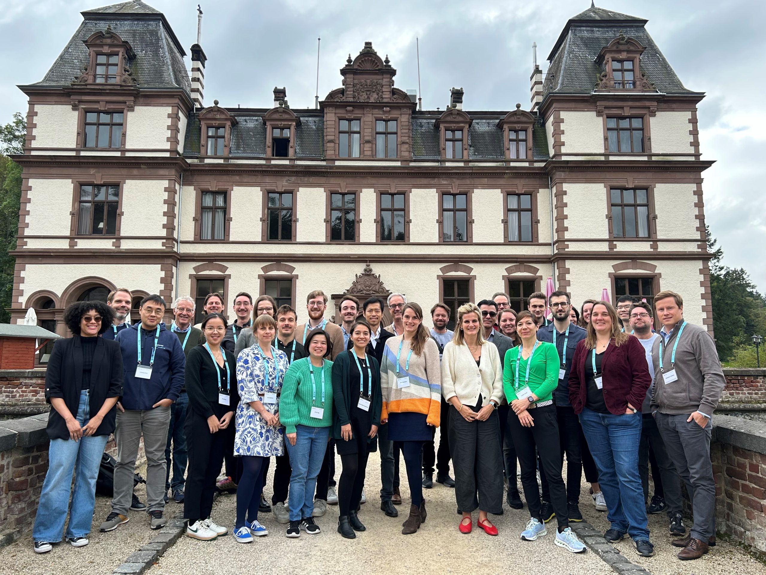 A group of around 35 people are standing in front of a large white stately home (Schloss Ahrenthal in Sinzig, Germany). They are all wearing blue lanyards. They are smiling at the camera.