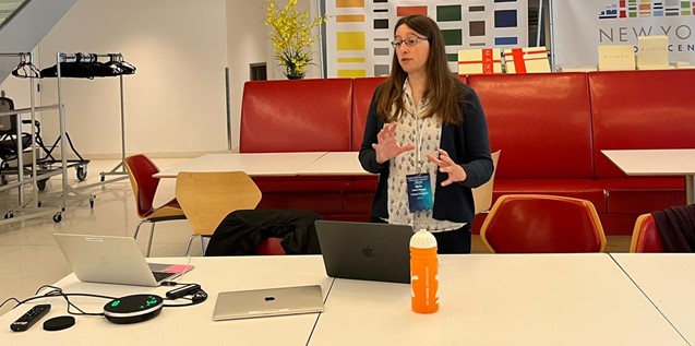 Marta is standing in front of two open laptops and is giving a presentation. Marta is a white woman with long brown hair and glasses, and is wearing a white patterned shirt and black cardigan. 