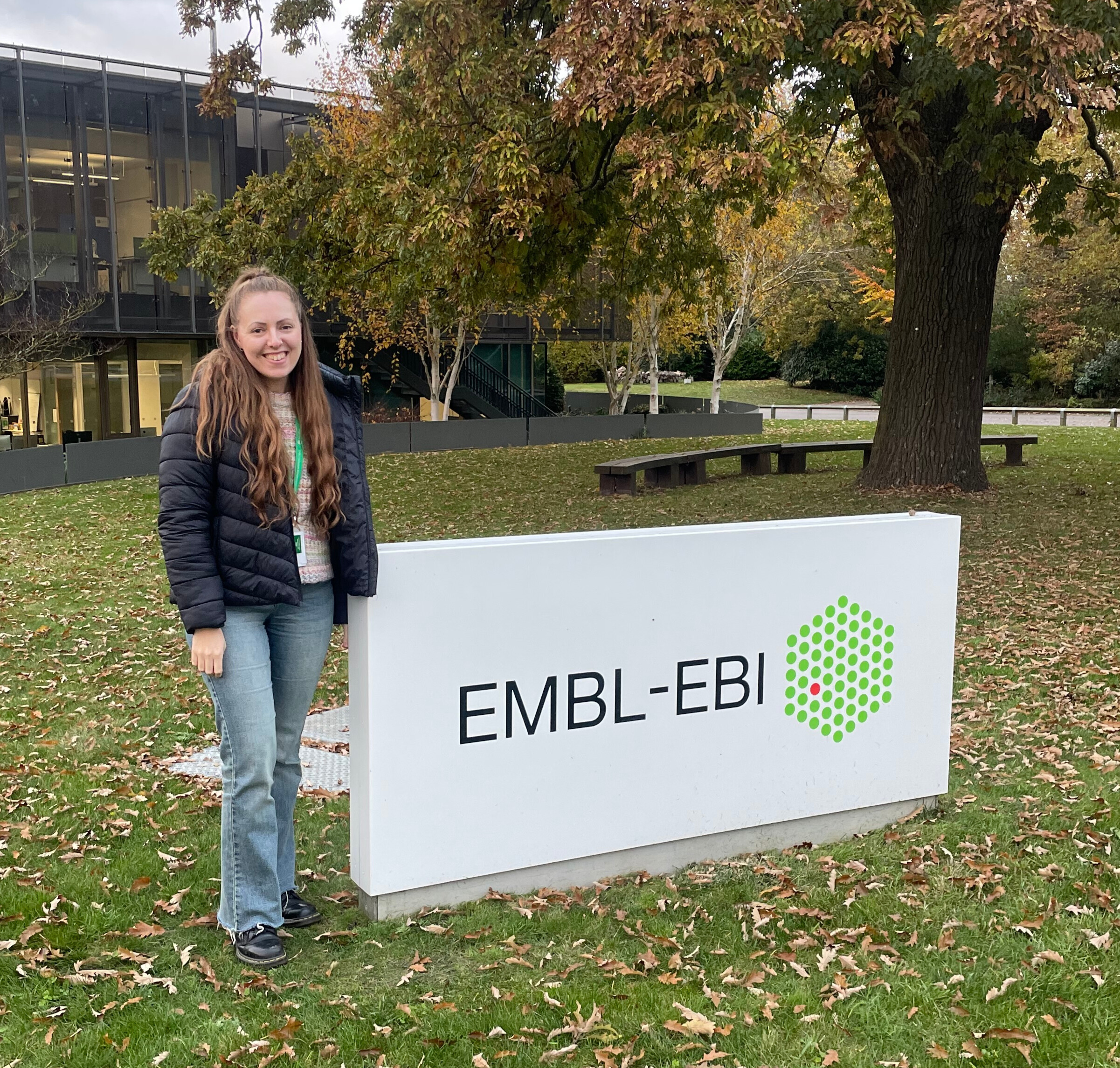 Xena is a young white woman with long brown hair, a black jacket and blue jeans. She is standing in front of a large white sign that says "EMBL-EBI" followed by the EBI logo (a green hexagon)