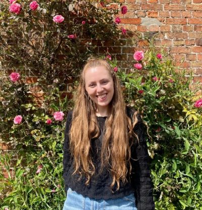 Xena is a young white woman with long brown hair. She is wearing a black top and blue jeans. She is smiling at the camera and standing in front of a wall with pink roses growing up it.