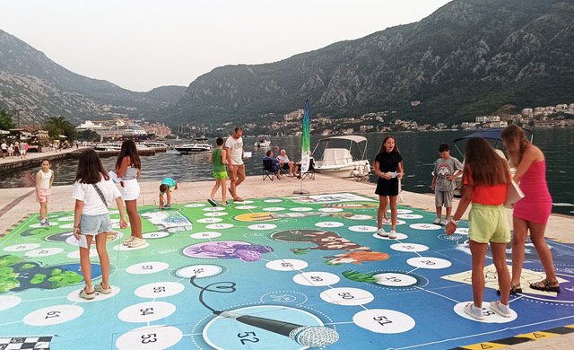 Close to the sea, a group of young people and families play a life-size game using dice.
