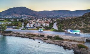 Two trucks with EMBL and TREC logos drive along a road next to the seaside, in the background are hills and houses from the suburbs of Athens.