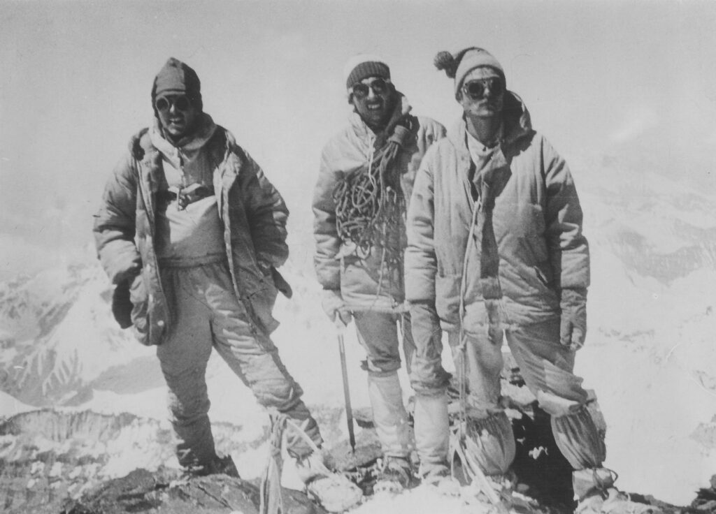 Black and white photo of three men wearing mountaineering gear, with mountainous peaks visible in the background. 