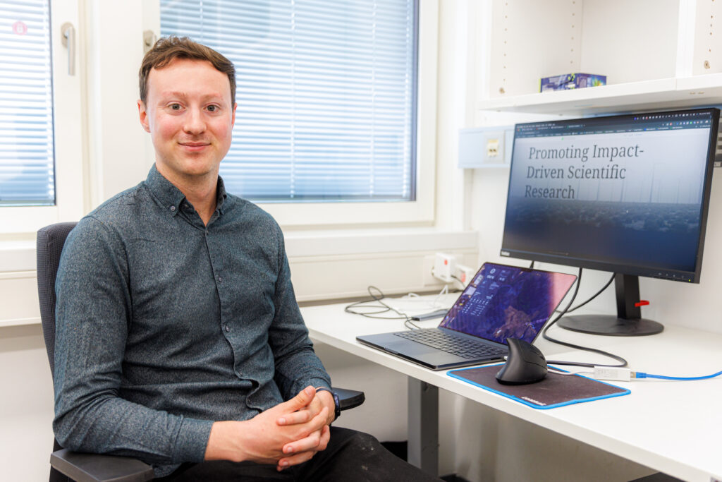 Male scientist at a desk, with a computer screen displaying the words 'Promoting Impact-Driven Scientific Research'