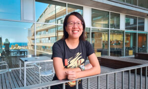 Portrait of a person on a terrace, with crossed arms leaning on the balcony rail. In the background the sea is reflected on the windows of the building.