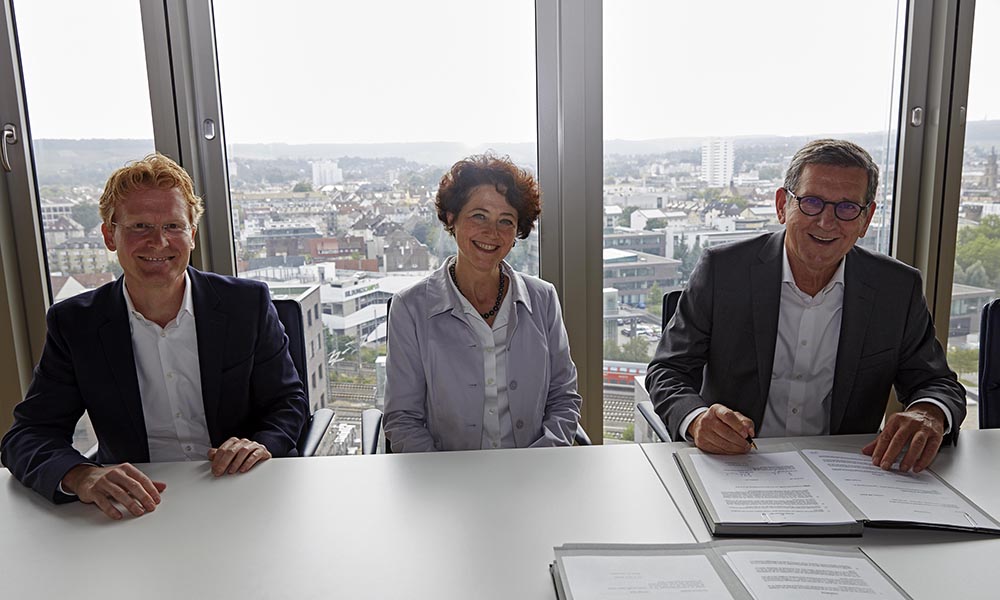 Three people seated at a table in front of a window with cityscape in background and table in foreground.