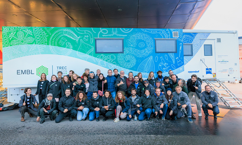 Around 40 international people of all ages posing together before a big mobile laboratory truck with logos of EMBL, TREC, and industry sponsors printed on its side.