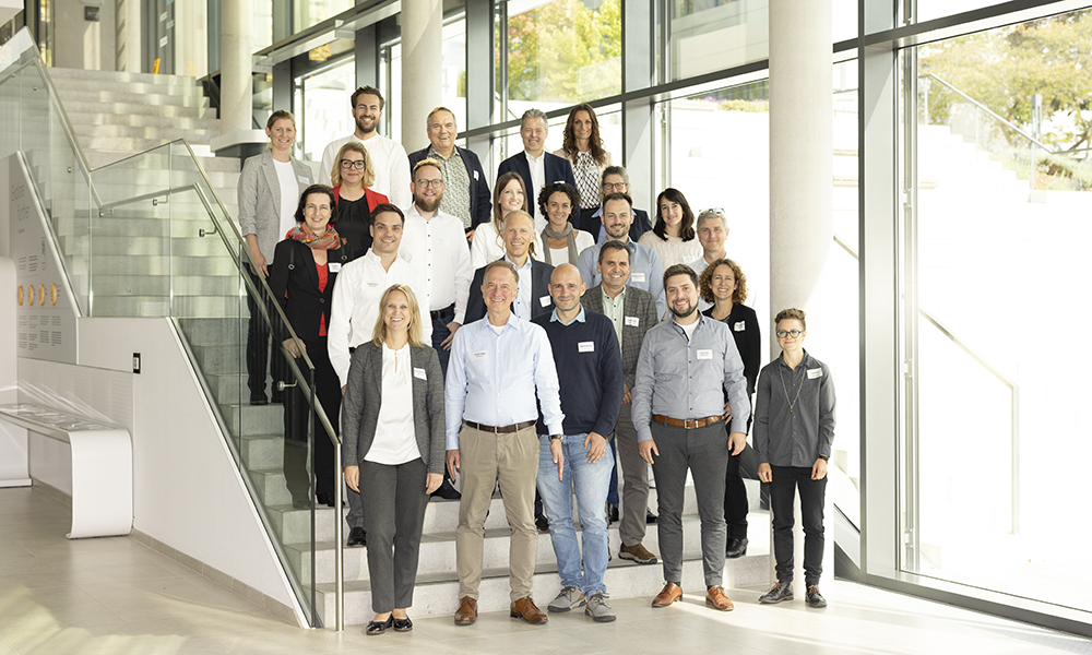 A group of about 20 people looking at the camera, standing on a set of stair inside a building with glass walls.