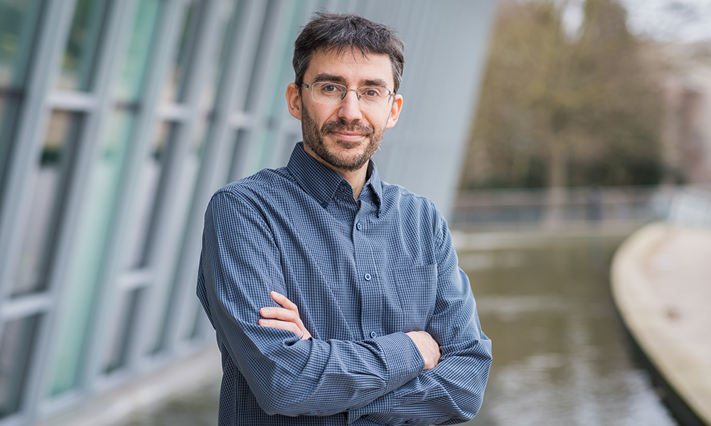 Man standing with arms crossed, wearing grey shirt and glasses.