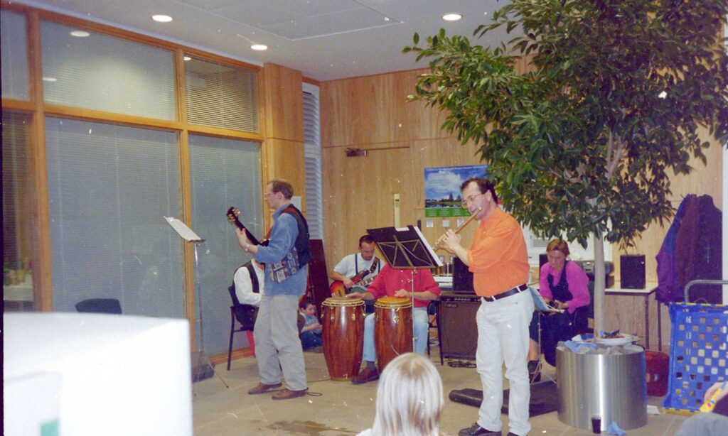 Band of musicians, including two guitarists, a flute player and a bongo drum player, jamming in an office building hallway.