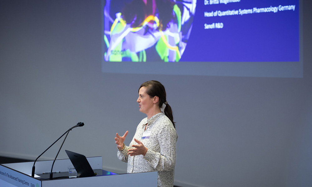 Woman talking at podium in front of large powerpoint slide projected on wall behind her