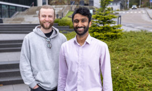 Two young males smiling to the camera with a mix of garden and buildings in the background.